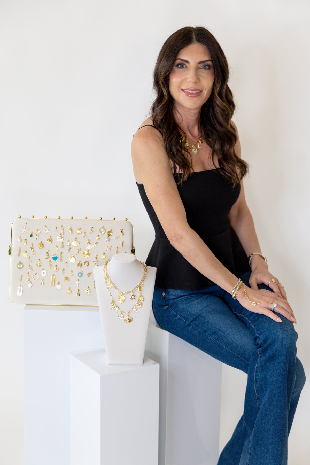 Lauren of Hustle and Heart sits on a white pedestal next to a white clutch bag and a white jewelry bust displaying gold  charm necklaces and charm bracelets.